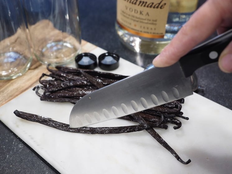 A sharp knife being held with tip in middle of vanilla bean slicing down the middle. Jars and vodka in background. www.thefreshcooky.com