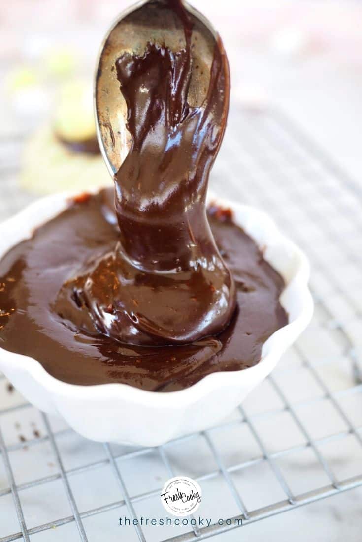 White bowl of chocolate ganache with spoon scooping some in, sitting on a cooling rack. 