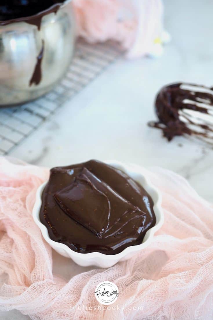 Ribbons of chocolate ganache in a small scalloped white bowl with whisk in background and pink cheesecloth around it. 