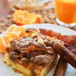 Overnight Cinnamon french toast slice on a plate with oranges, maple syrup, sausage and bacon.