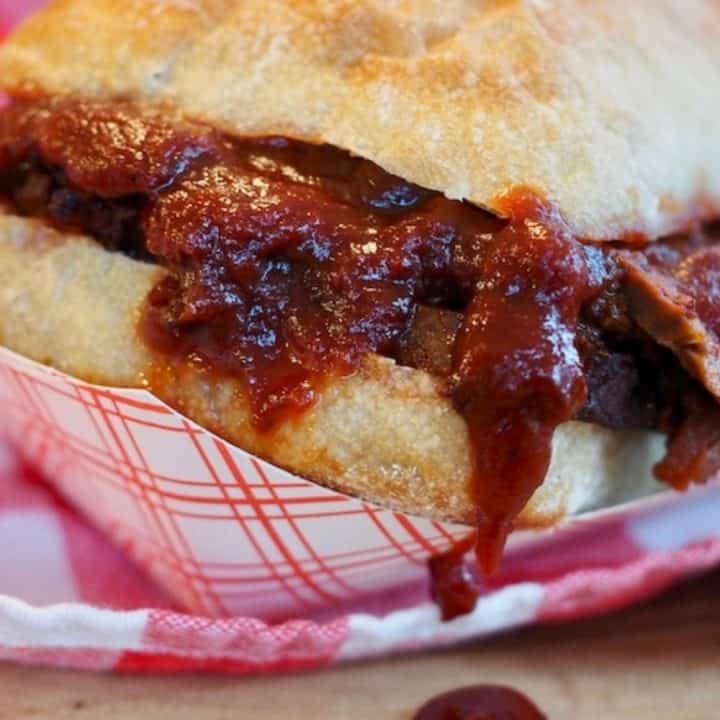 Pinterest Image ciabatta roll, stuffed with barbecue beef brisket sandwich with a drip of bbq sauce on wooden board. sitting on red and white checked napkin.