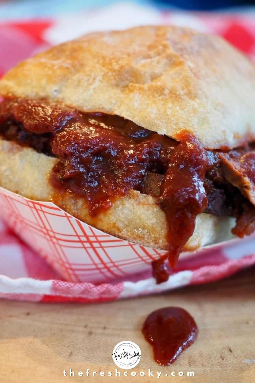 Pinterest Image ciabatta roll, stuffed with barbecue beef brisket sandwich with a drip of bbq sauce on wooden board. sitting on red and white checked napkin.