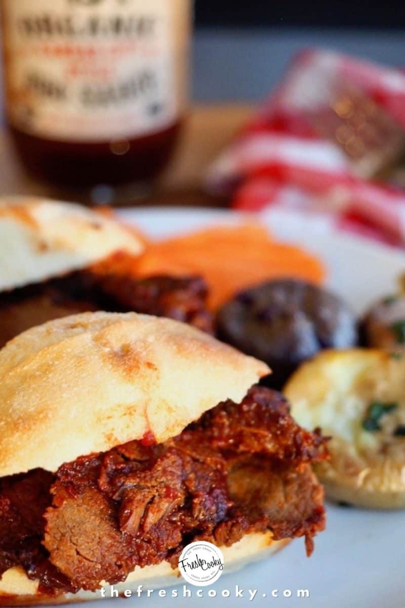 barbecue beef brisket sandwich on plate with smashed potatoes and carrots with bottle of BBQ sauce in background. 