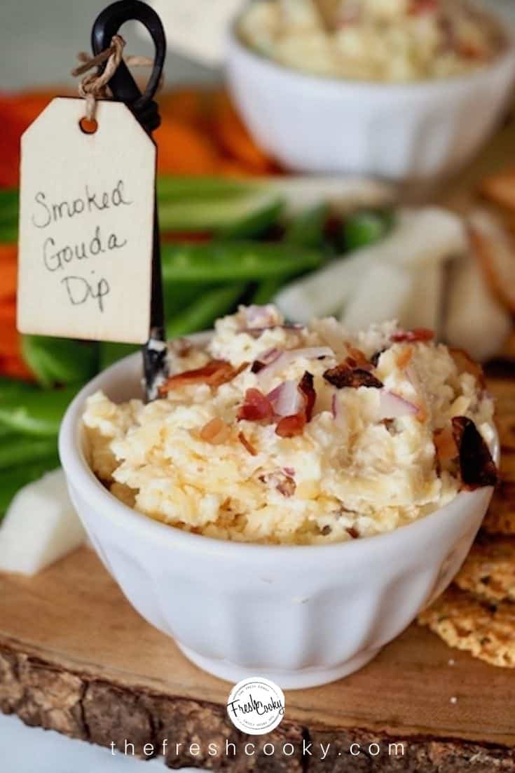 Smoked gouda cheese dip in white serving bowl with crackers and veggies nearby. 