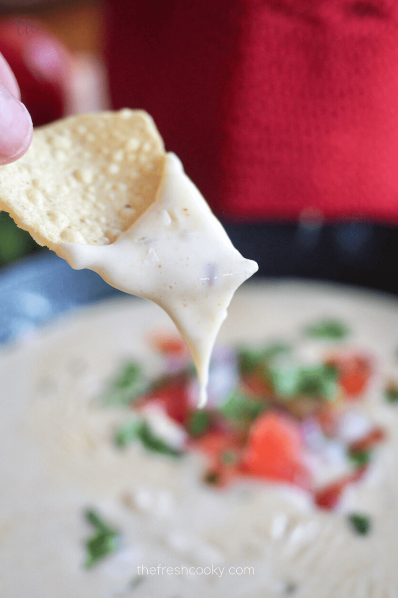 Hand holding tortilla chip dipping in Mexican White Cheese Queso Dip.