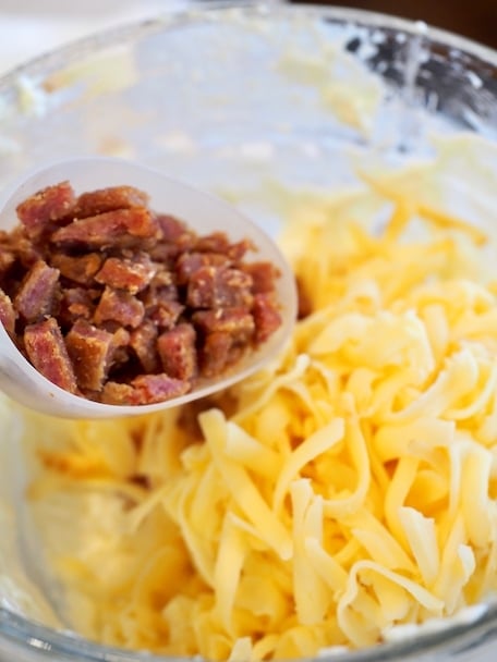 Bacon Bits in a measuring cup over Shredded Smoked Gouda cheese in a larger glass mixing bowl for smoked gouda cheese dip.