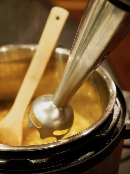 Immersion blender going into Instant pot with wooden spoon in background.