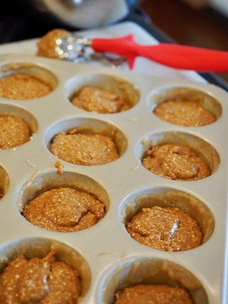 Muffin Tin with bran muffin mixture spooned into each tin cup, red handled scoop in background. 
