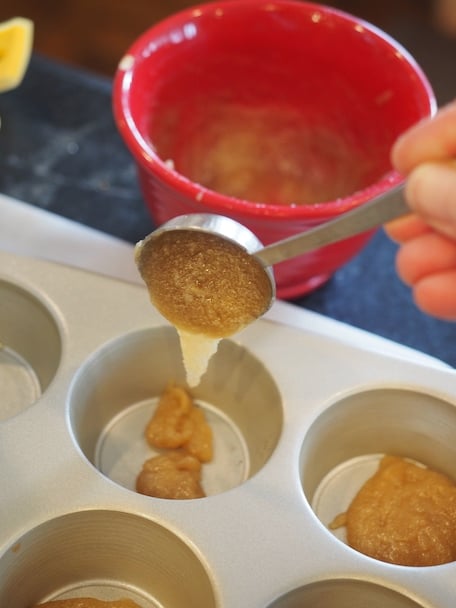 A tablespoon filled with glaze being poured into cups of muffin tins with red bowl in background.