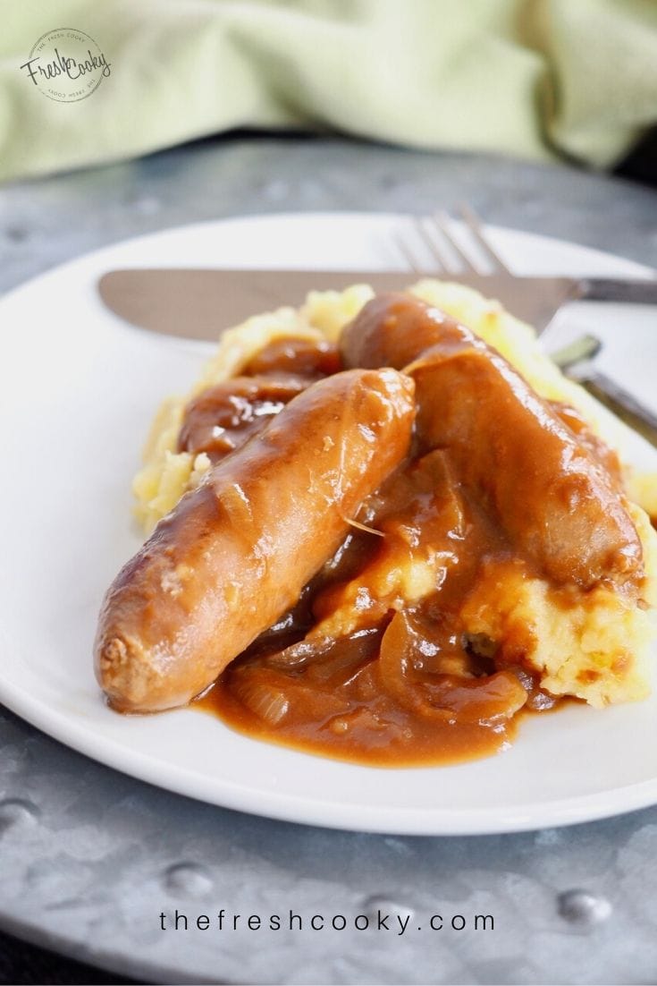 Plate piled high with two bangers (sausages) smothered in gravy on top of a pile of mashed potatoes. 
