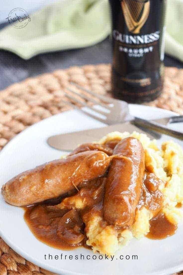 White plate with rustic knife and fork behind with two bangers (sausages) on top of a mound of fluffy mashed potatoes, smothered in guinness stout gravy.