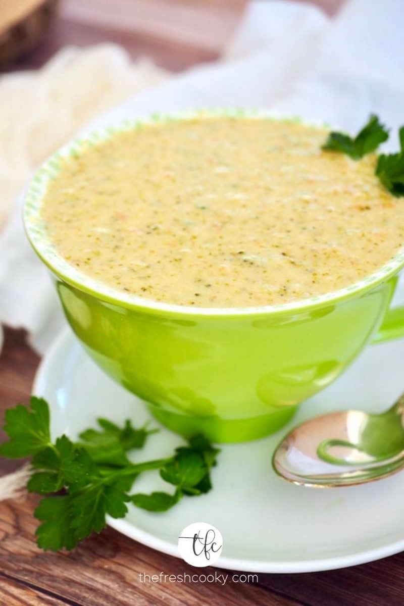 Bright green bowl filled with creamy broccoli cheese panera soup with spoon and sprigs of parsley laying around.