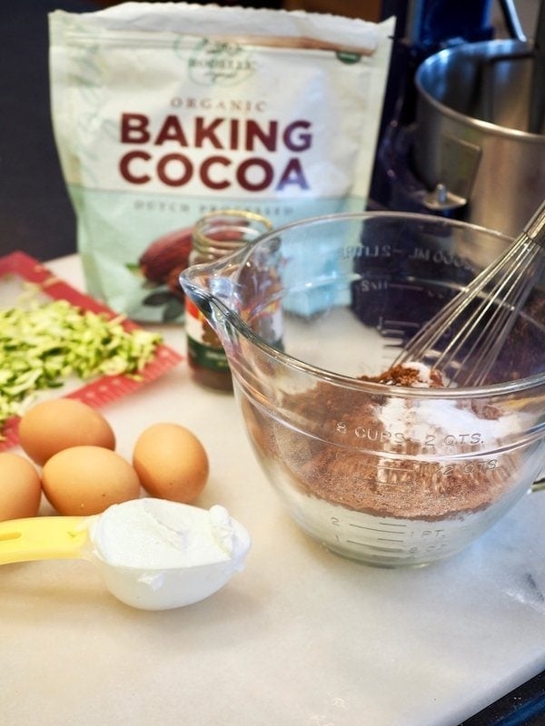 chocolate_cake_ingredients sitting on marble board. Left to right. Zucchini, baking cocoa, espresso powder, 4 brown eggs, a glass mixing bowl with flour, cocoa powder, salt, leavening and a whisk, greek yogurt. www.thefreshcooky.com