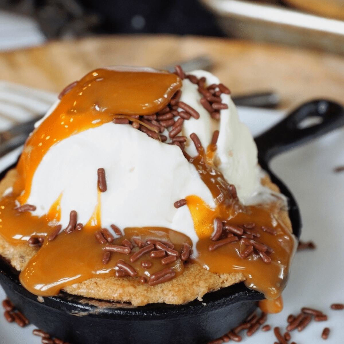 Half baked cookie square image of cookie in mini cast iron skillet with vanilla ice cream, caramel sauce and chocolate sprinkles.