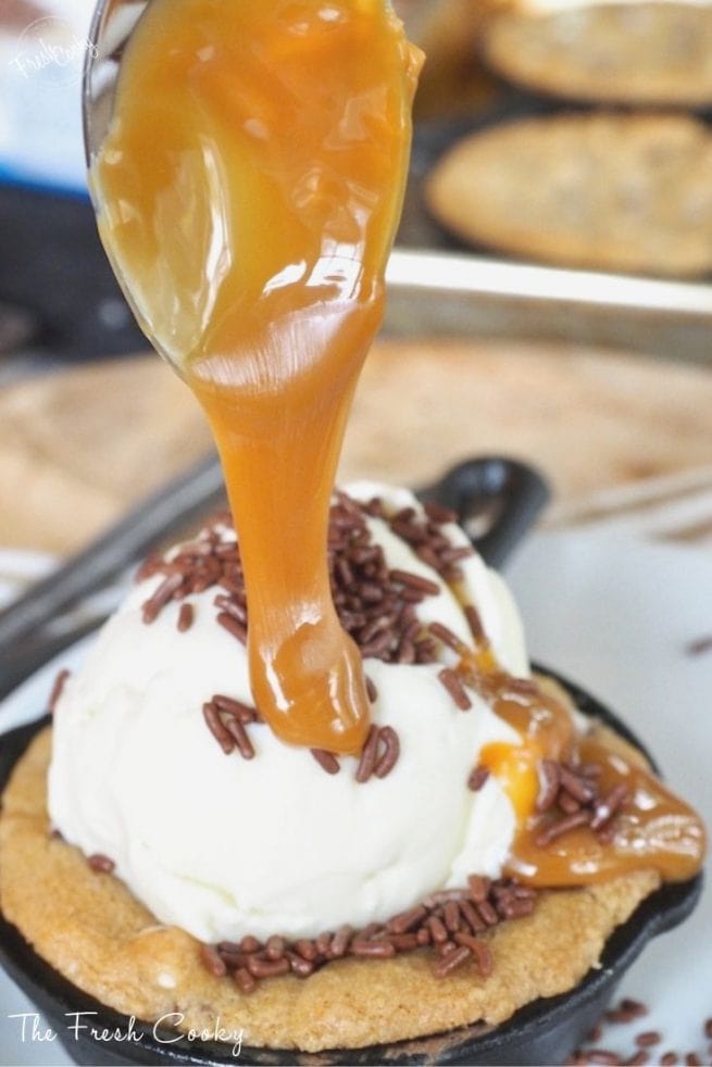 Gooey half baked chocolate chip skillet cookie with scoop of ice cream and a spoon drizzling caramel sauce on top.