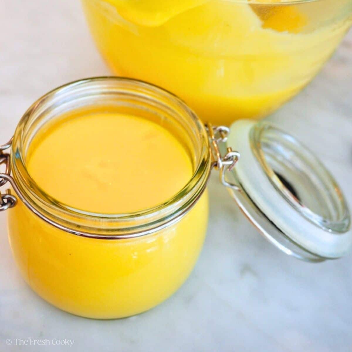 Lemon curd in a jar with a bowl behind, creamy, bright yellow, tangy lemon curd.