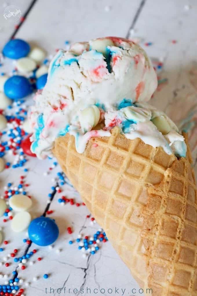 Waffle cone with bright red, white and blue ice cream scoop in the cones with non-pariels and M&M's laying around it. 