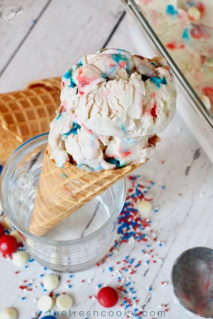 Two scoops of red, white and blue no churn patriotic ice cream in a waffle cone with sprinkles and jimmies laying around. 