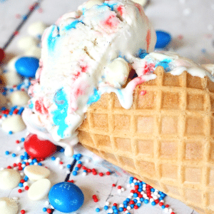 Red white and blue ice cream scooped into a waffle cone with red, blue m&m's and white chocolate chips sprinkled around.