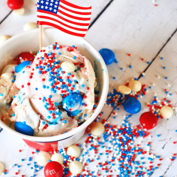 Red, white and blue ice cream scoop in a patriotic cup with an AMerican flag and sprinkled with red, white and blue m & m's.