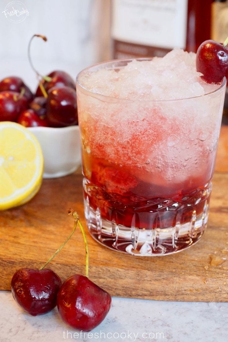 Image of a fizzy Cherry Bomb Cocktail on wooden cutting board with fresh cherries and lemon.