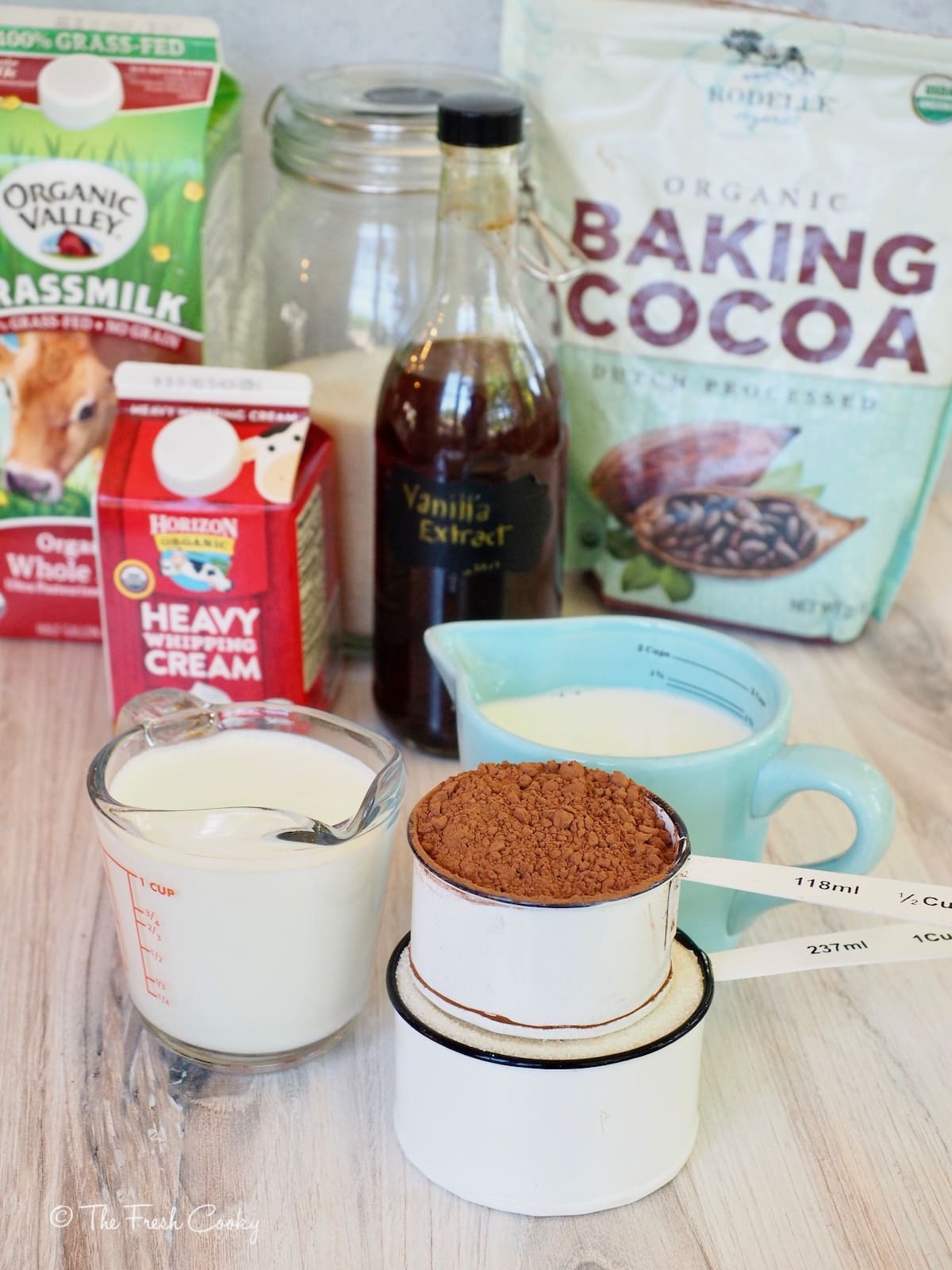 Ingredients on wooden board for chocolate ice cream. Left to right. Grassfed whole milk, all natural sugar, Rodelle Baking Cocoa, Vanilla Extract, Heavy Cream and each item measuring in a measuring cup.