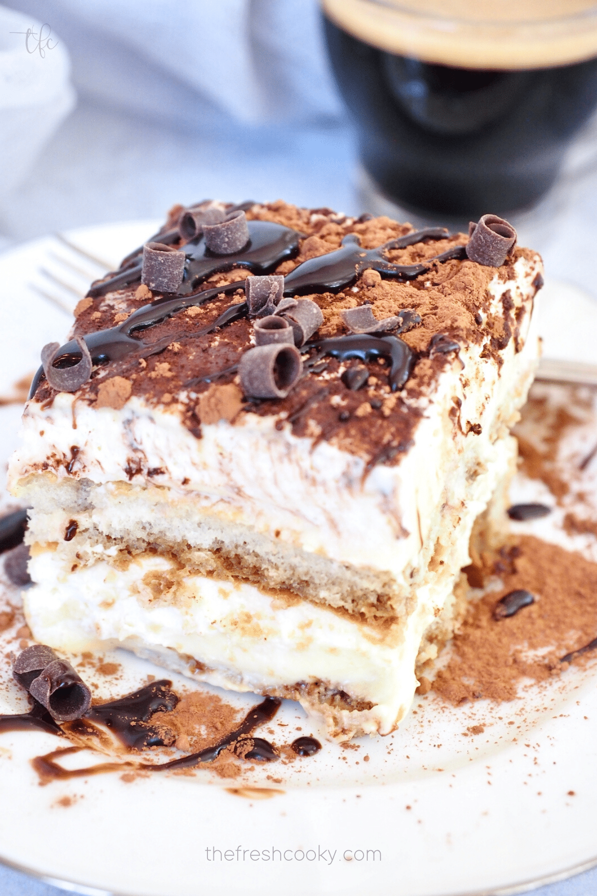 Traditional Italian Tiramisu Recipe on a plate dusted with cocoa powder, drizzled with chocolate and an espresso in the background.