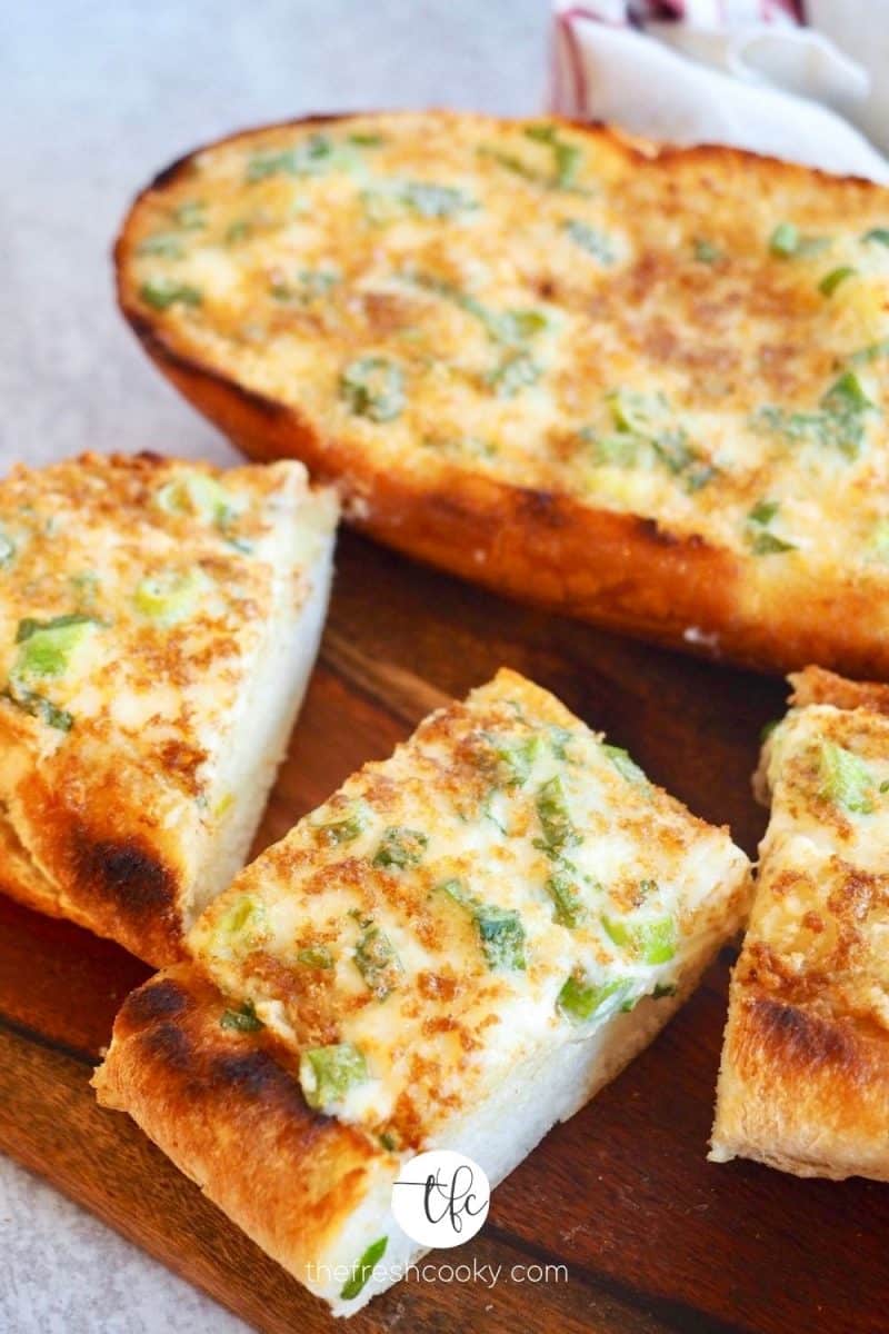 Image of cheesey garlic bread on cutting board with half loaf and several slices in forefront.