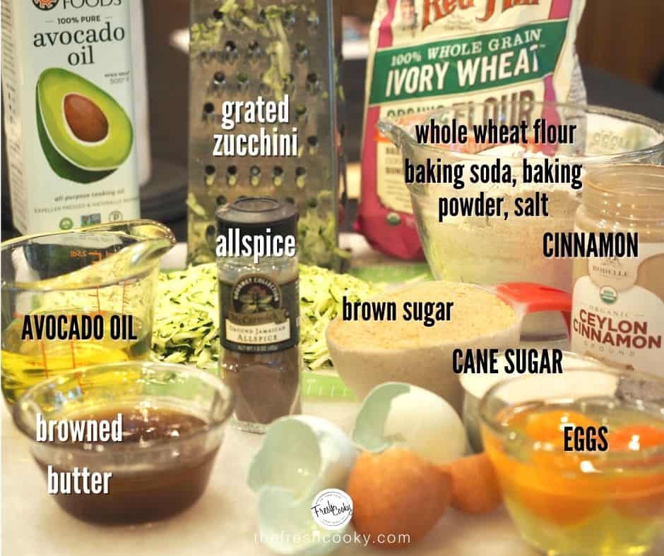 ingredients for snickerdoodle zucchini bread, left to right. Avocado Oil, grater with grated zucchini, ivory wheat flour, glass mixing bowl with flour, baking soda, baking powder and salt, cinnamon in a jar. front row browned butter in small glass bowl, jar of allspice, measuring cup with brown sugar, smaller measuring cup with cane sugar and glass bowl with eggs, cracked egg shells in front.