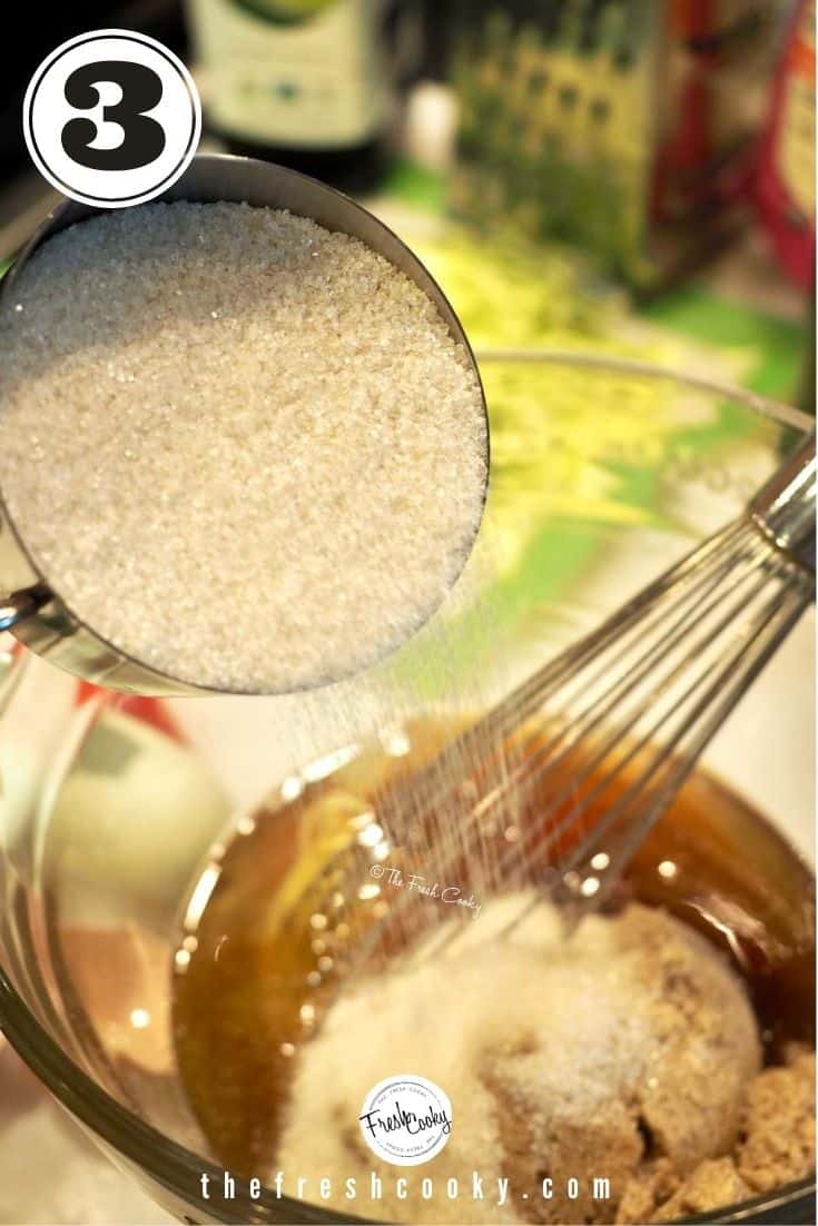 glass mixing bowl, with metal cup of natural cane sugar pouring into bowl filled with oil, browned butter and a whisk