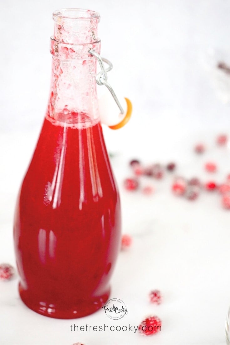 image of bottle of bright red cranberry simple syrup with swing top lid and cranberries around it.