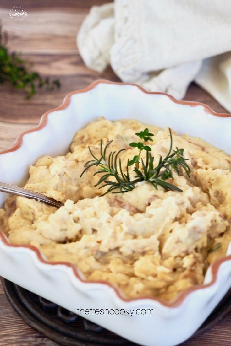 Rustic mashed potatoes in white clay square baking dish with rosemary and thyme in the middle and thyme in background along with natural colored napkin. 