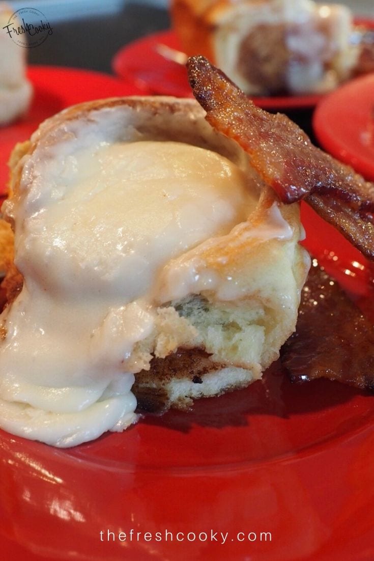 Pin with image of frosted cinnamon roll on red plate with candied bacon and oozing Cinnabon frosting a cream cheese frosting.