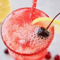 cranberry spritzer bubbling in glass with red and white polka dot straw, garnished with lemon and cherry.