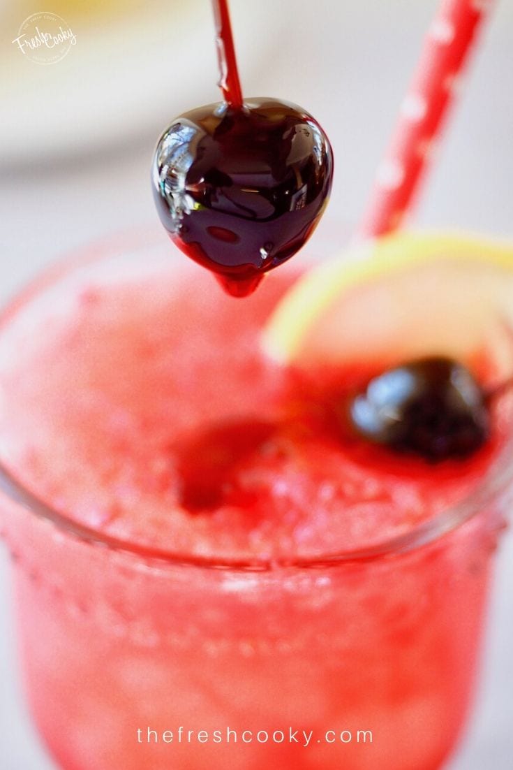 Bright rich red glossy candied cherry in syrup being placed into cranberry spritzer glass.