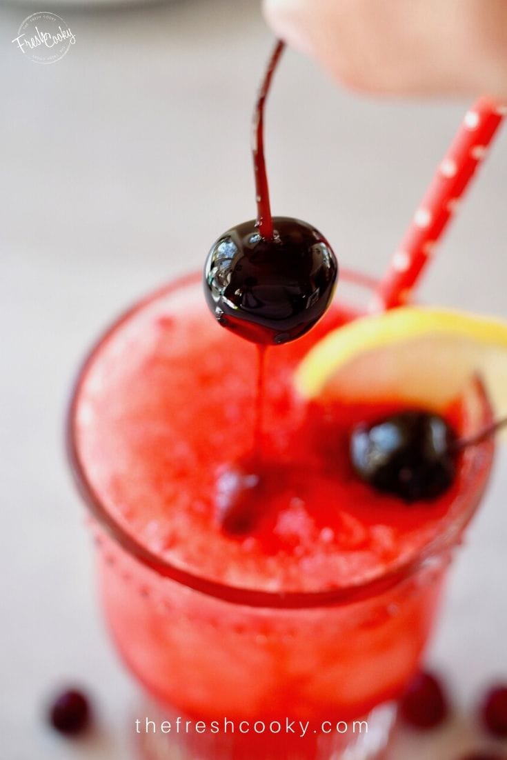 Top down image of glass of cranberry spritzer with a cherry dripping with juice being placed inside the bright red drink. 