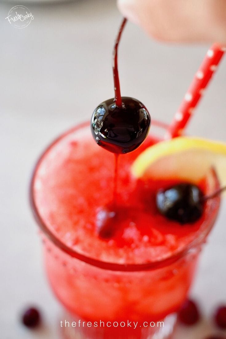 Top down image of glass of cranberry spritzer with a cherry dripping with juice being placed inside the bright red drink.