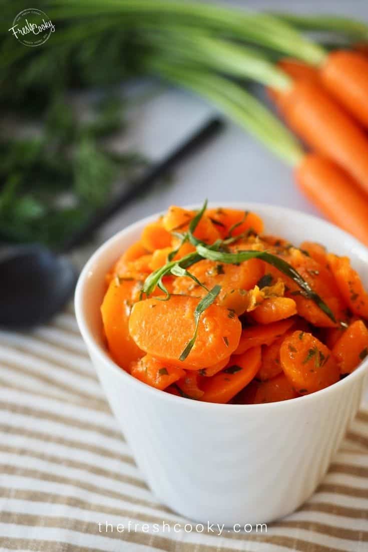 A white bowl filled with bright orange glazed tarragon carrots with fresh carrots in the  background. 