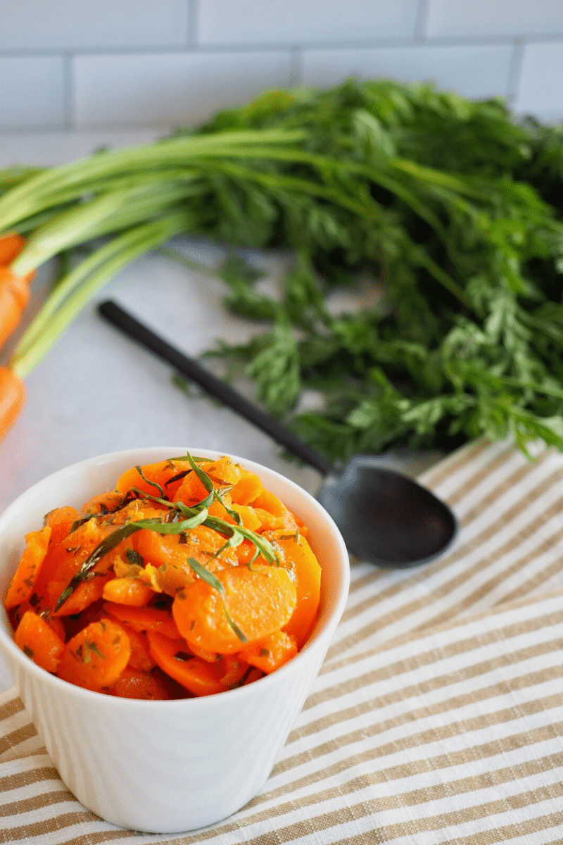 Bowl of glazed instant pot carrots on striped tea towel with fresh carrots and a serving spoon.