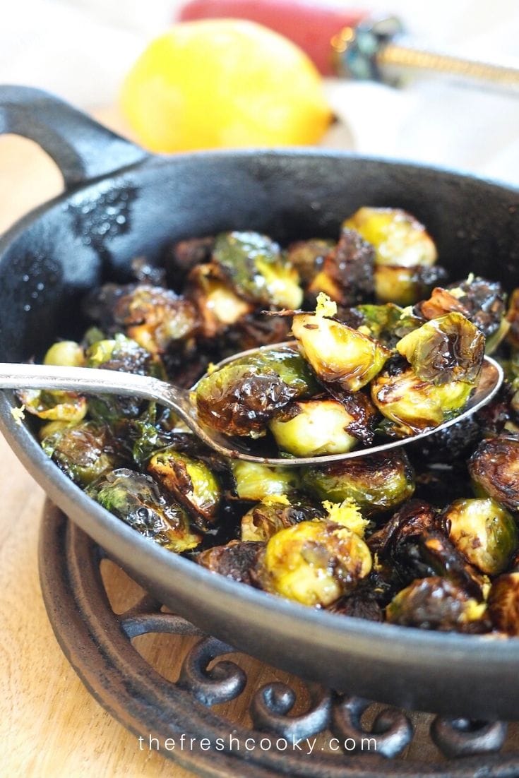 Spoonful of roasted crispy brussels sprouts with honey balsamic glaze in a cast iron pan, with zester and lemon in background. 