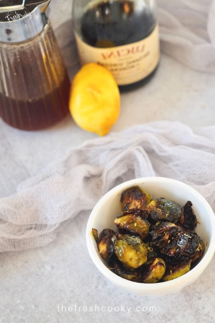 Small white bowl of roasted crispy brussels sprouts on table with honey, lemon and balsamic vinegar in background. 