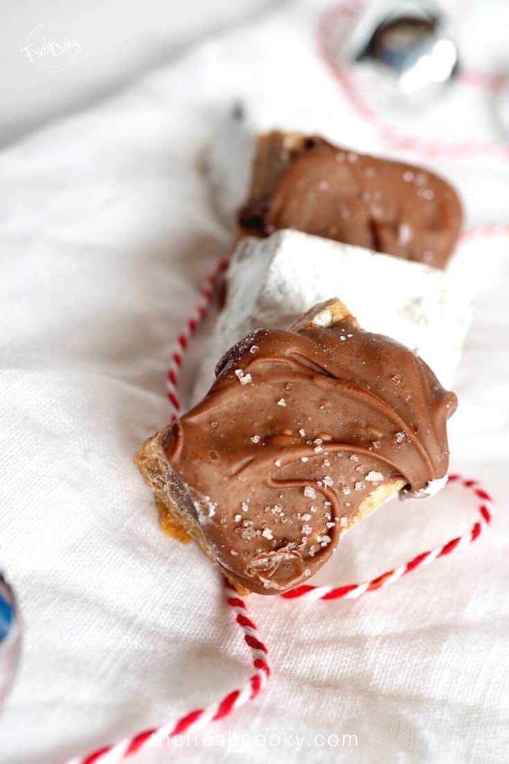 Salted chocolate graham bite with red and white bakers twine on white napkin with jingle bell in background.