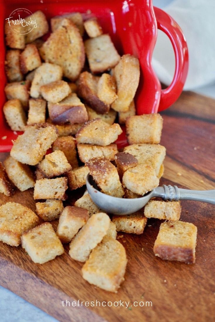 Croutons in red ceramic bowl spilled onto wooden cutting board with pewter spoon. 