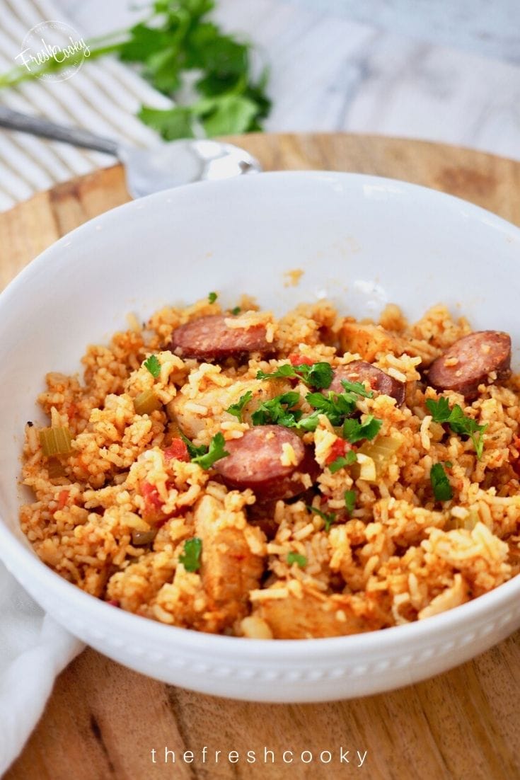 White bowl filled with jambalaya, spoon in background on wooden charger with parsley laid on top.