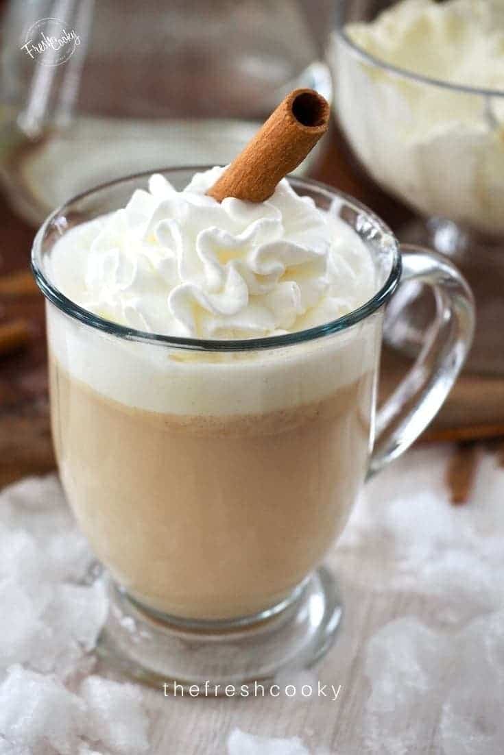 Glass mug filled with creamy hot buttered rum drink topped with whipped cream and a cinnamon stick. 