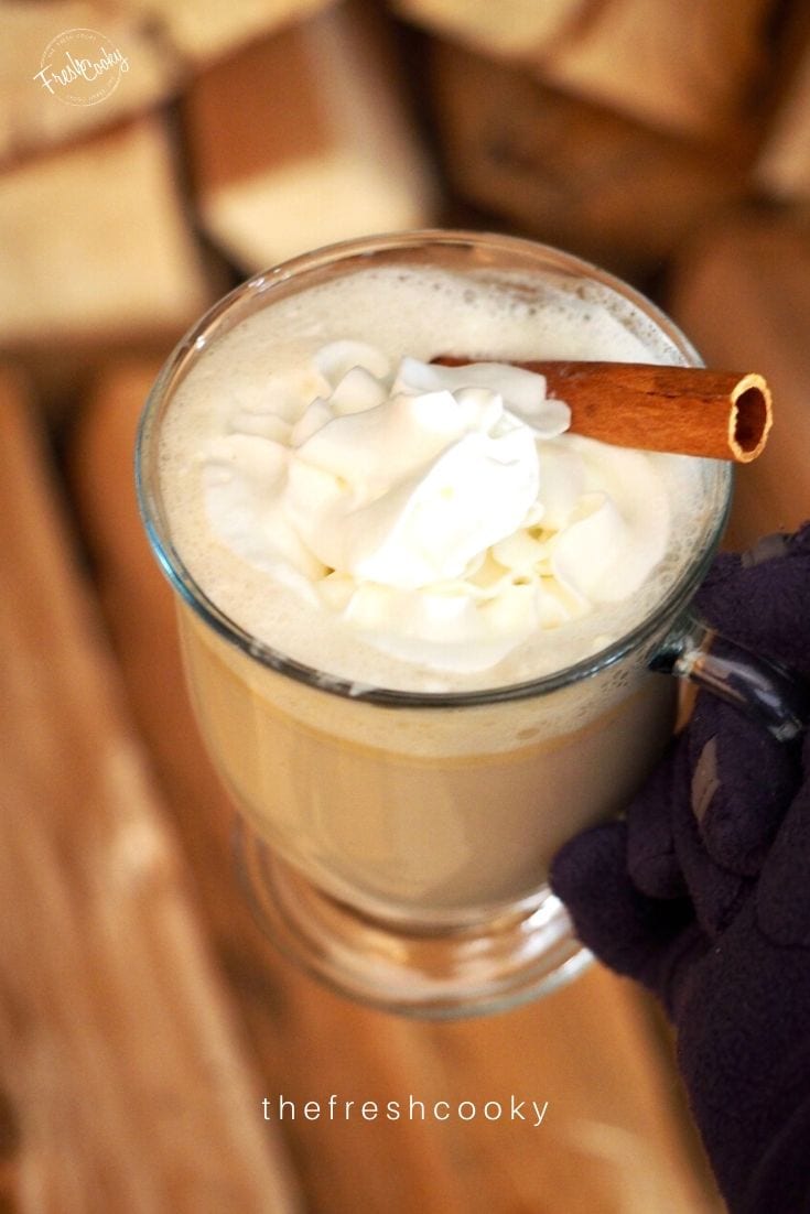 Gloved hand holding glass mug filled with hot buttered rum topped with whipped cream and cinnamon. 