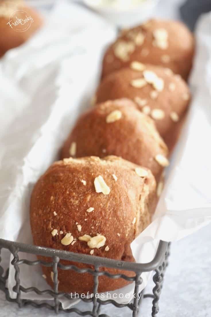 Long basket of sweet brown bread molasses rolls topped with oats in a parchment lined wire basket. 