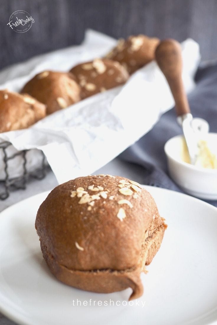 Pinterest image of sweet molasses brown bread rolls on a white plate with a basket of rolls in the background, butter and butter knife included. 