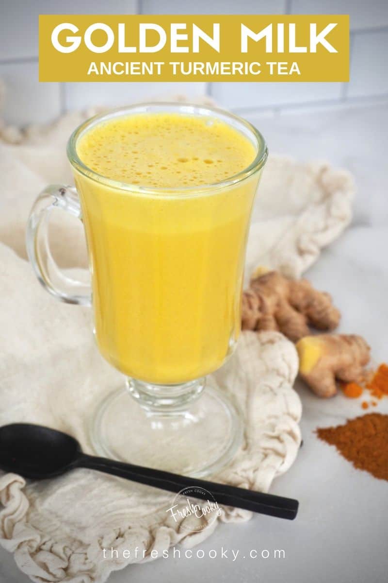 Glass mug filled with bright yellow golden milk or turmeric tea on a rustic napkin with a black spoon, ginger root and other spices in background. 