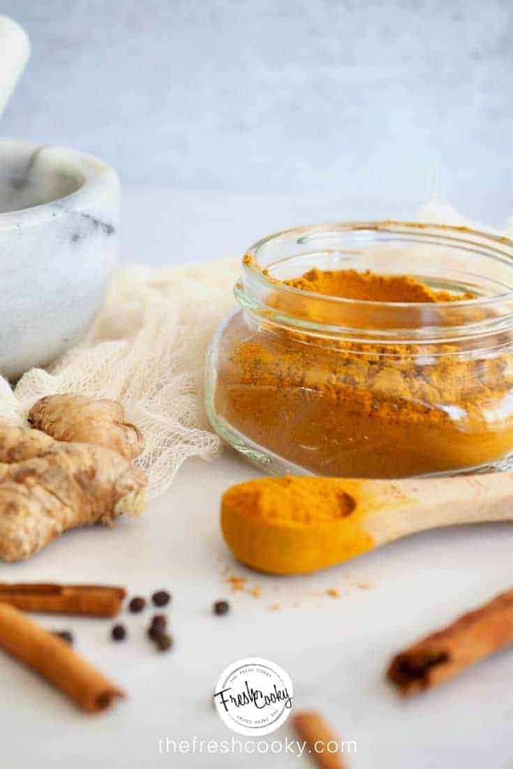 golden milk spice mix with mortar and pestle in background as well as fresh ginger root, black peppercorns and cinnamon sticks, with jar filled with golden milk spice mix